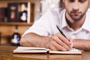 Confident bar owner. Close-up of handsome young male bartender in white shirt leaning at the bar counter and writing something in note pad